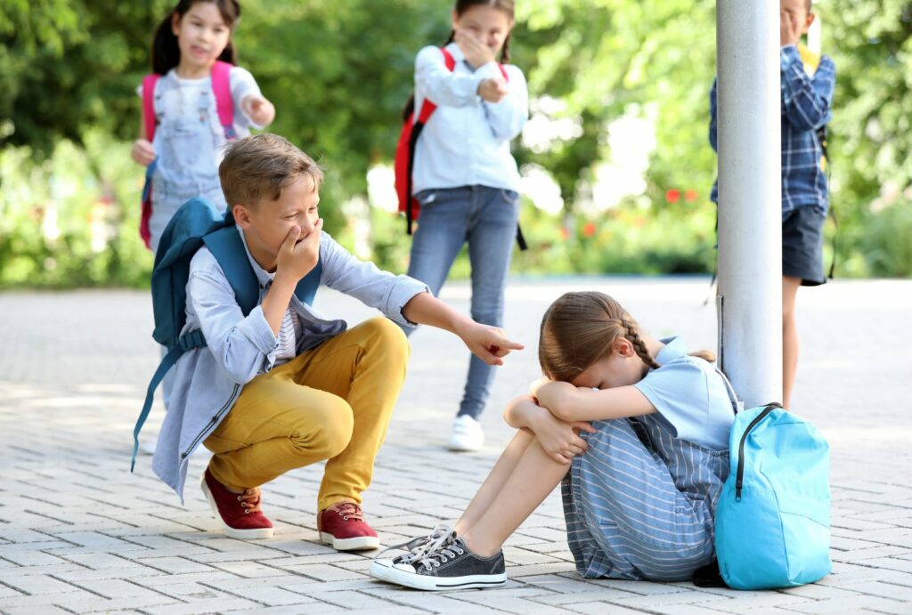 Dzieci śmieją się z dziewczynki siedzącej skulonej na podwórku.